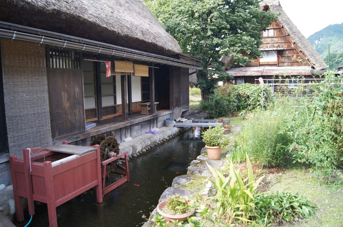 A traditional thatched farmhouse stands beside a garden and water channel in Shirakawa-go.
