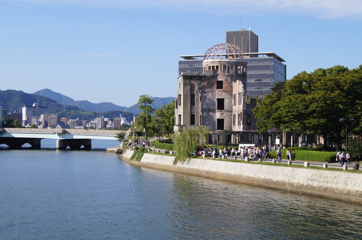 The Atomic Bomb Dome stands beside the river with Hiroshima buildings and mountains behind it.