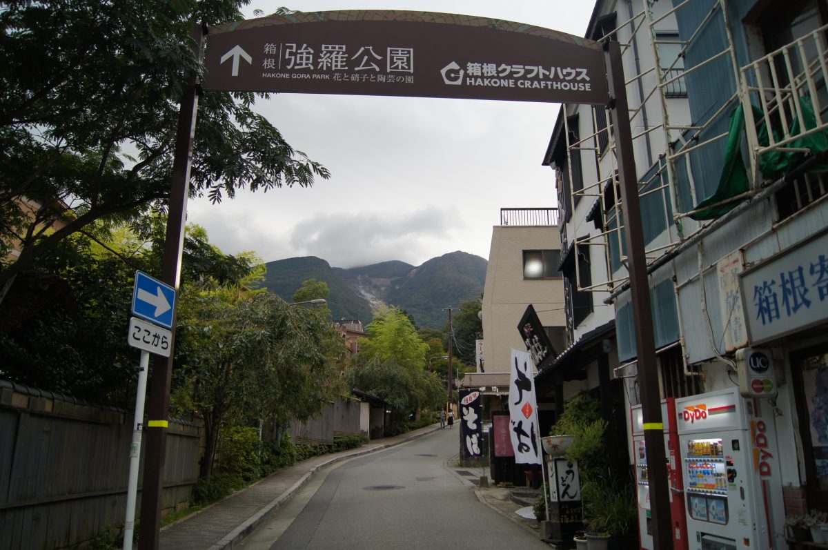 Hakone 98 A street beneath a hanging sign with wooded mountains rising behind it in Hakone.