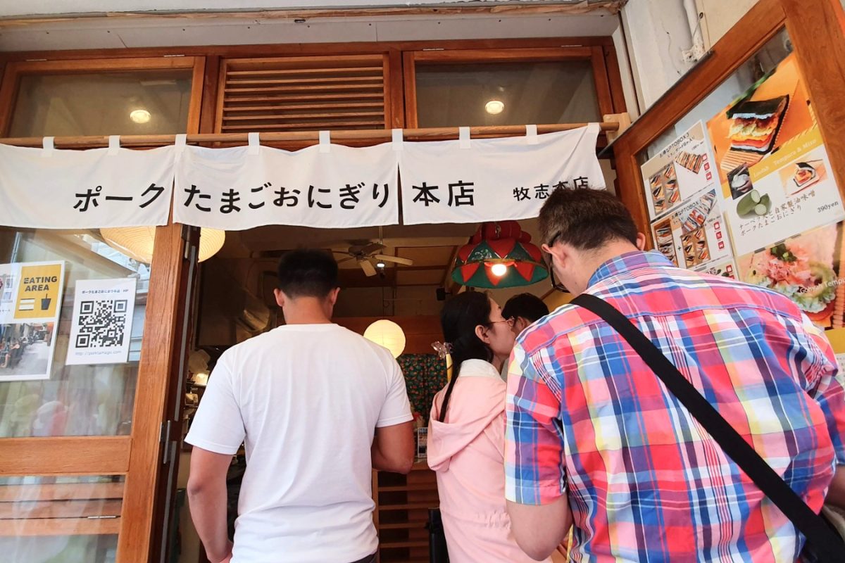 People queuing at a rice sandwich shop beneath a white Japanese sign.