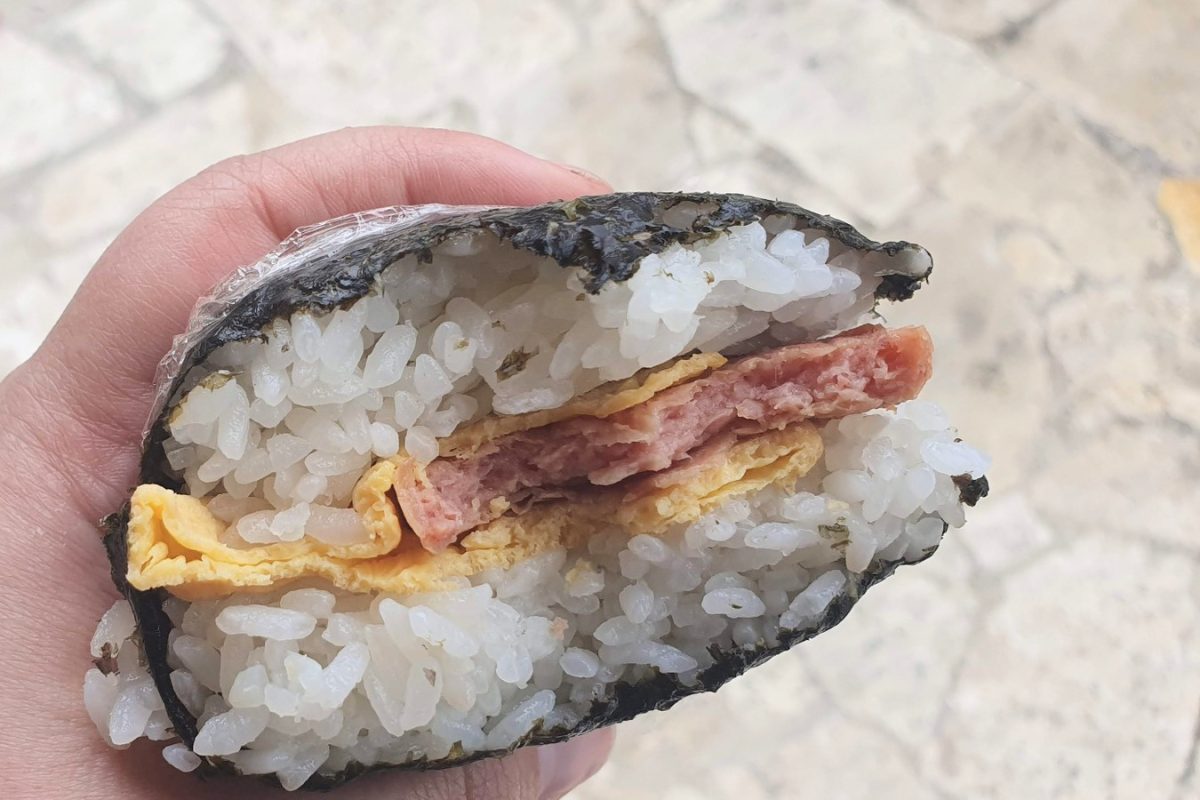 Hand holding a seaweed-wrapped rice sandwich filled with pork and egg.