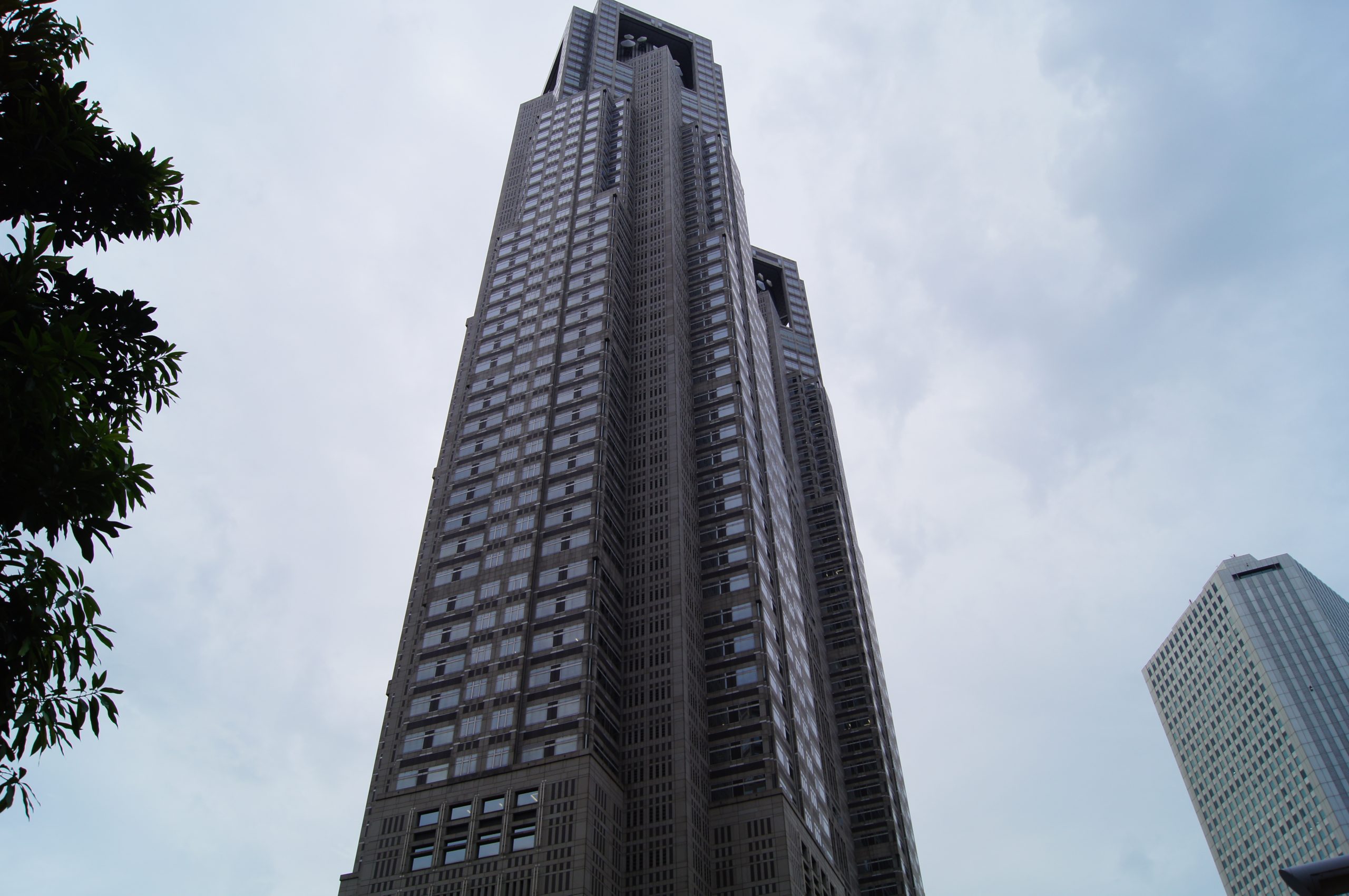 Tokyo Government Building 12 scaled Grey gridded towers with stepped upper levels against a cloudy sky, with foliage on the left and another tower on the right.