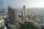 A stepped-roof high-rise among rectangular towers and roads, with dense buildings stretching into the distance beneath a pale hazy sky.