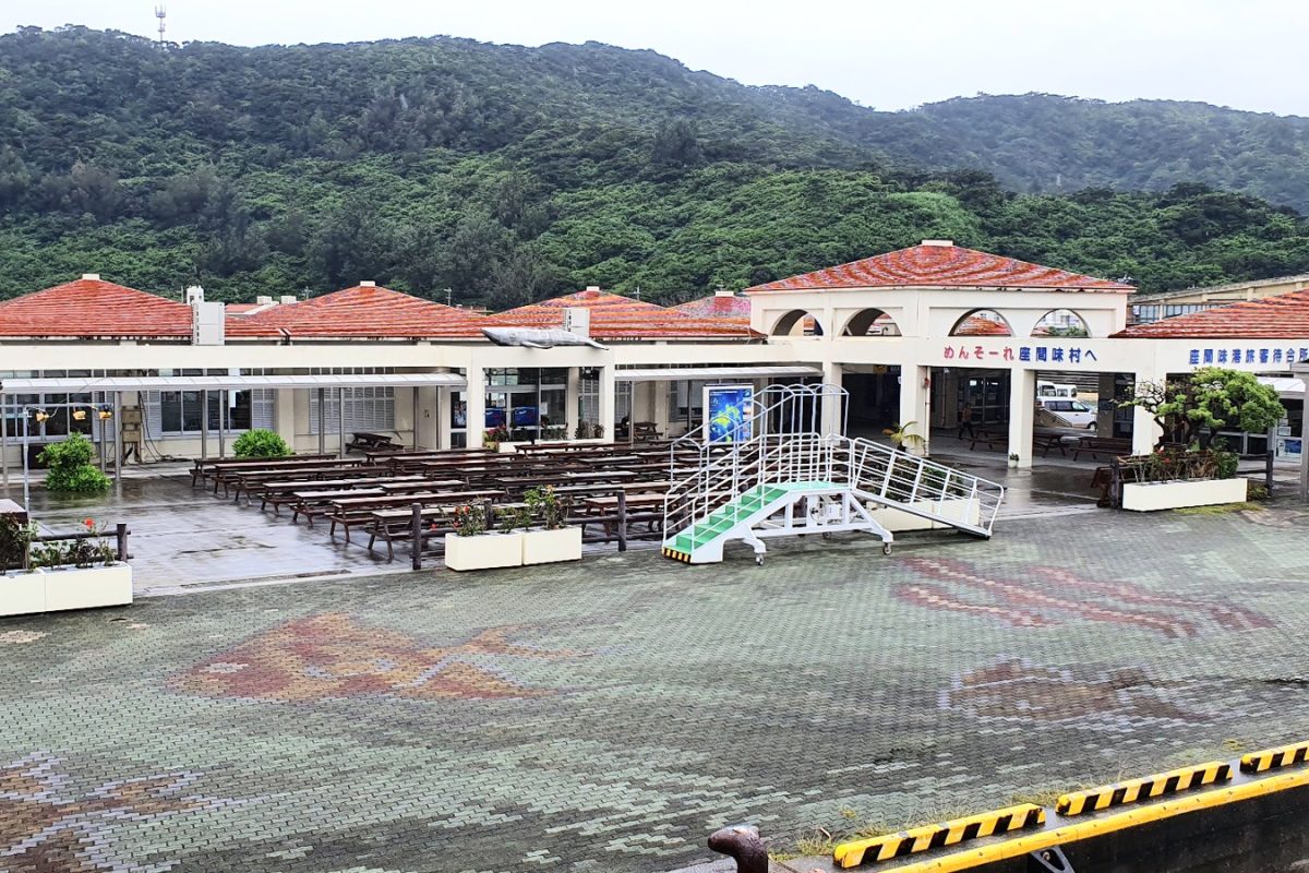 Zamami ferry terminal with red tiled roofs, seating and forested hills behind.