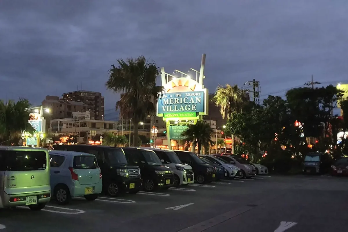 Illuminated American Village entrance sign beside palms and parked cars in May 2019